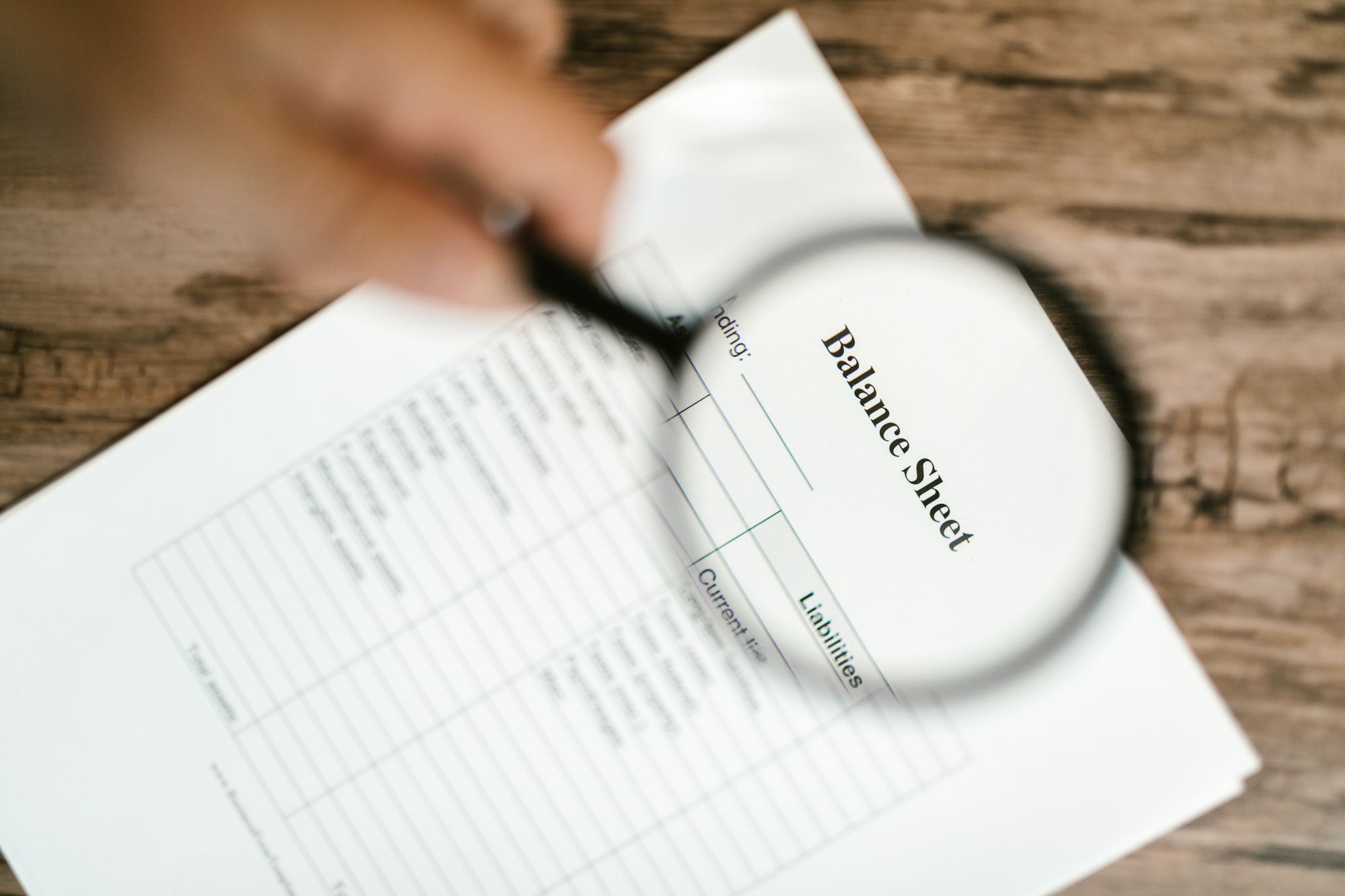 A magnifying glass held over a balance sheet showing assets and liabilities